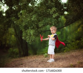 Little Blond Boy In Red Hero Cloak With Toy Catapult In Summer White Shorts And Sneakers Standing On A Forest Hill