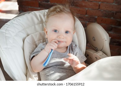 little blond boy sits in a children's high chair and nibbles a spoon