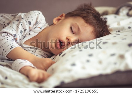 Little blond boy sleeping in his bed. Restless sleep of a baby with a cold. The kid sleeps with his mouth open. My little boy sleeping by the light of the window.