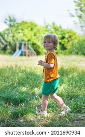 little blond boy smiling and running on green grass in summer against the backdrop of the playground