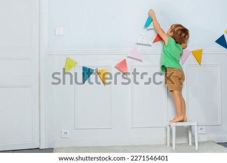 Little blond boy standing on the stool put colorful party pennants chain, garland with flags on a wall in living room