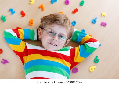 Little blond child with glasses playing with lots of colorful plastic digits or numbers, indoor. Kid boy wearing colorful shirt and having fun with learning math