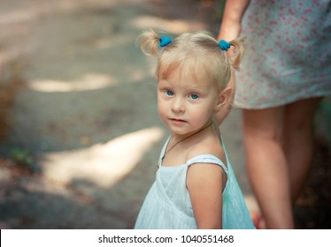 Little Blond Girl With Blue Eyes Holding Her Mother's Hand And Looking Into The Camera. Cute Baby Girl Two Years Old In A White Summer Dress.