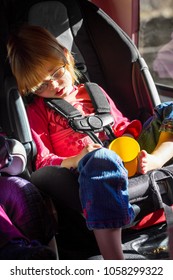 A Little, Blond Girl On A Roadtrip Has Fallen Asleep And Is Drooling In Her Carseat.  She Has An Empty Cup In Her Lap And There Are Crumbs Below Her, By Her Feet.