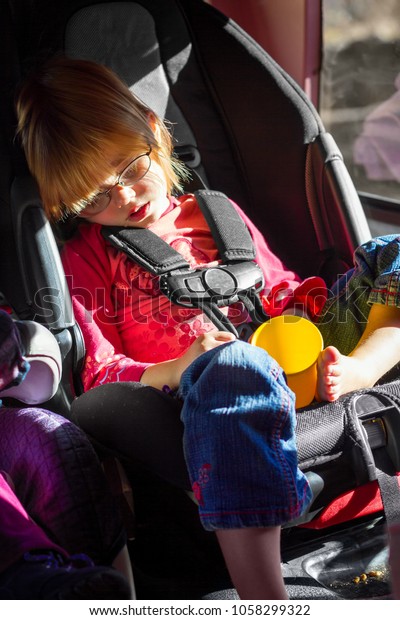 A little, blond girl on a\
roadtrip has fallen asleep and is drooling in her carseat.  she has\
an empty cup in her lap and there are crumbs below her, by her\
feet.
