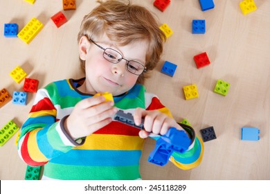 Little Blond Kid Boy Playing With Lots Of Colorful Plastic Blocks Indoor. Child Wearing Colorful Shirt And Glasses, Having Fun With Building And Creating.