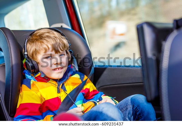 Little blond\
kid boy watching tv or dvd with headphones during long car driving\
on family vacations. Leisure for children for long drive. Preschool\
child sitting in safe car\
seat.