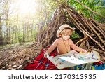 Little blond scout boy sit with treasury map playing treasure hunt game over hut of branches