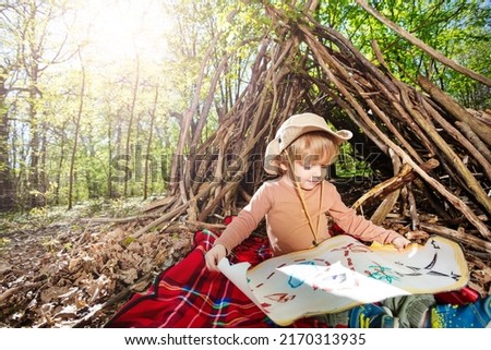 Little blond scout boy sit with treasury map playing treasure hunt game over hut of branches