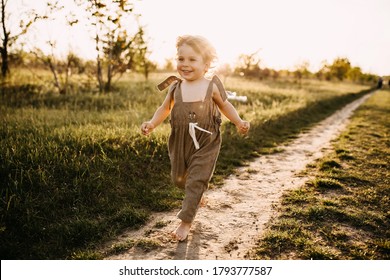Little blonde boy with curly hair, wearing vintage jumpsuit, running on a dirt path, barefoot, at sunset.