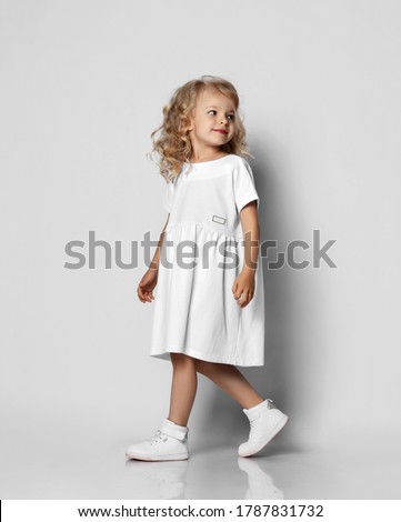 Little blonde curly positive princess girl in white casual dress and sneakers standing walking with curly hair over grey wall background. Stylish comfortable everyday fashion for children concept