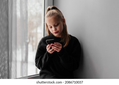 Little Blonde Girl In Black Sweater Sitting On Window Sill And Using Smartphone. Bored Child Writing On Phone Indoors. Sad Kid Home Alone During Lockdown. Texting, Chatting, Reading.