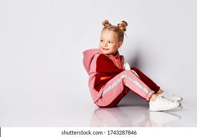 Little blonde girl with buns hairstyle, in colorful tracksuit, sneakers. She laughing, sitting on floor, posing isolated on white. Childhood, fashion, advertising and sport. Close up, copy space - Shutterstock ID 1743287846