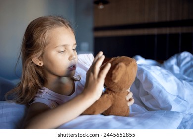 A little blonde girl in cute pink pajamas is playing with a plush brown teddy bear on a large bed in an amazingly beautiful hotel room