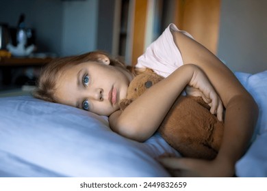 A little blonde girl in cute pink pajamas is gently hugging her plush brown teddy bear on a large bed in a cozy and modern beautiful hotel room