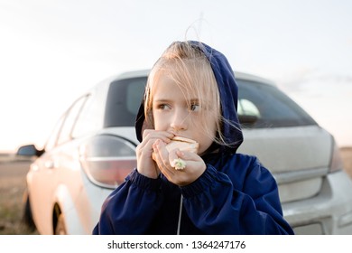 Little Blonde Girl Eating A Sandwich On A Walk Near A White Car On The Background Of The Sunset In The Summer