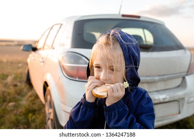 Little Blonde Girl Eating A Sandwich On A Walk Near A White Car On The Background Of The Sunset In The Summer