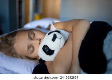 A little blonde girl in an embrace with a teddy bear panda sleeps on a large and cozy bed in a delightfully cozy hotel room