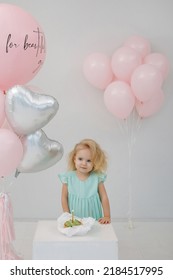Little blonde girl in a green dress blows a candle on a birthday cake in the studio with pink and silver heart-shaped balloons. Festive decoration. Valentine's Day.