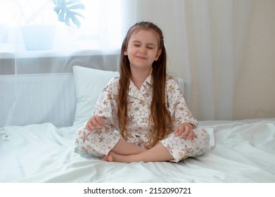 Little blonde girl with long hair in pajamas is doing yoga exercise in sunny room. A child does breathing exercises on white bed.