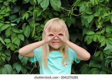 A little blonde girl makes glasses or a batman mask on her face with her hands. The child makes faces against the background of green leaves. 