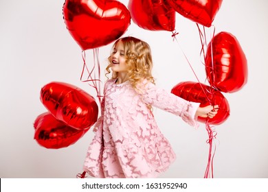 little blonde girl in a pink dress is smiling and holding a lot of red heart-shaped balloons, space for text. the concept of Valentine's daylittle blonde girl in a pink dress is smiling and holding a 