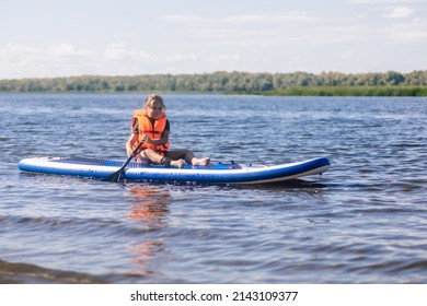 Little blonde girl puddle boarding on lovely lake rowing with oar in hands looking at rippled water in vest life jacket. Active holidays. Inculcation of love for sports from childhood. - Shutterstock ID 2143109377