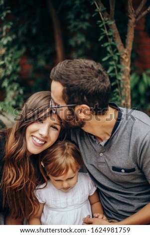 Little blonde girl sitting between mon and dad. Father kisses mother on the head. Daughter enjoying with her young parents in the garden. Love and family concept.