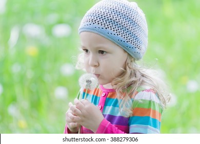 Little Blonde Girl Wearing Striped Knitted Hat Blowing On White Puffy Dandelion Seed Head At Her Hand