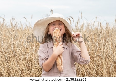 Little blonde girl in a white hat laughs and eats a French bread baguette. A happy child in a rye field in summer