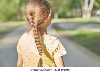 A Little Blonde Girl In A Yellow Casual Dress With A Pigtail On Her Head, Standing With Her Back To The Camera. High Quality Photo