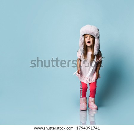 Little blonde kid with long hair, dressed in pink blouse, plush hat, pants and boots. She raised her hands, looking confused, posing on blue background. Fashion, advertising. Full length, copy space