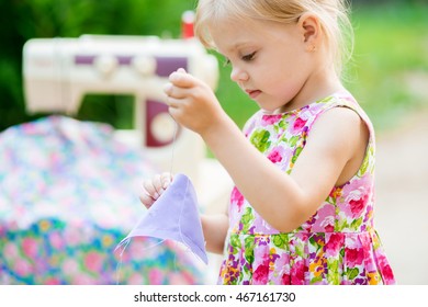 little blonde smiling lovely child 4 years old (girl) in cotton dress sewing needle (hand) of the purple cloth on a background of a sewing machine with a cotton cloth beautiful outdoors - Powered by Shutterstock