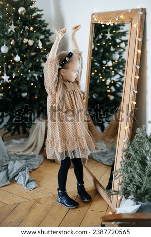 Little blondhaired girl in a party dress dancing in front of the mirror with a decorated and lighted Christmas tree in the background 