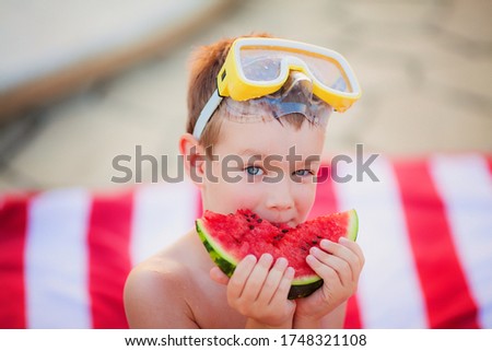 Little blue-eyed blond boy wearing yellow Diving Goggles eating watermelon sitting on the striped red and white beach towel in the summer