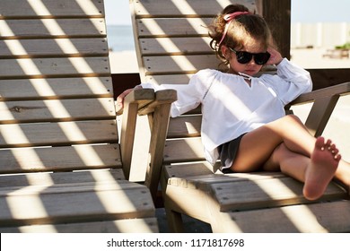 Little Bossy Girl Wearing Black Sunglasses Sitting On A Wooden Chair On The Beach