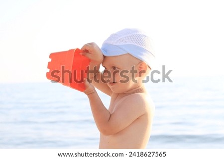 A little boy, 1 year old, plays on the sea with a toy plastic red bucket. A boy pours sand from a bucket onto the sea on the beach. In the background is the sea or ocean. 