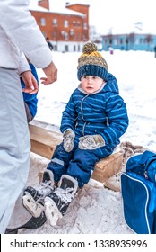 Little Boy 2-4 Years Old, Sitting On A Bench For Shoes, In The Winter In A City Park On A Snow-covered Skating Rink. In Blue Overalls And Black Skates.