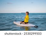 Little boy 9-10 years old paddling on stand up board having fun during summer beach vacation. Kid paddle surf in summer beach. Child on a SUP or Stand Up Paddle board in tropical beach, turquoise sea.