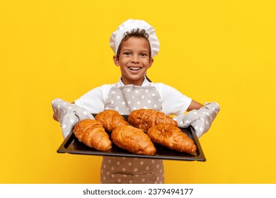 little boy african american baker in uniform and gloves holding baking sheet of fresh croissants on yellow isolated background, child cook offering pastries and bread - Powered by Shutterstock