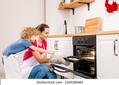 Little Boy In Apron Pointing At Lamp In The Oven While Standing By His Mom Holding Tray With Raw Cookies