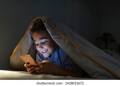 Little Boy In Bed Under A Blanket Looking At The Smartphone At Night. Teen Boy Lying On Front In The Bed And Using Smartphone. 7 Years Old Child Boy Using Smartphone At Night In Bed