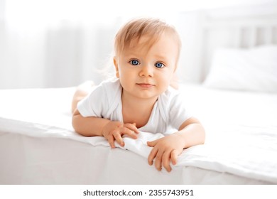 a little boy, a blond-haired child with blue eyes in a white bodysuit, is on all fours at home in the bedroom on a bed with white linens and looks at the camera - Powered by Shutterstock