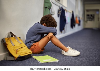 Little boy with books sitting alone in school hallway while crying. African american boy feeling depressed after bullying by classmates. Sad school boy at school after a bad grade on the class test. - Powered by Shutterstock