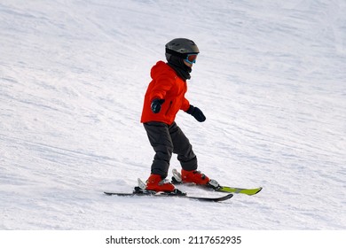 A little boy in a bright red jacket and boots is skiing down the mountainside by himself. He maintains balance during the downward movement by balancing with his hands