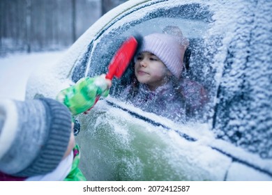 Little Boy Brushes Snow From A Car Window, Into Which Girl Looks Out. It Looks As If A Boy Is Cleaning A Girl, Funny Moment. Family Road Trip, Active Weekend In Any Weather, Winter Car Safety