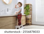 Little boy brushing teeth while standing on step stool near bathroom vanity indoors