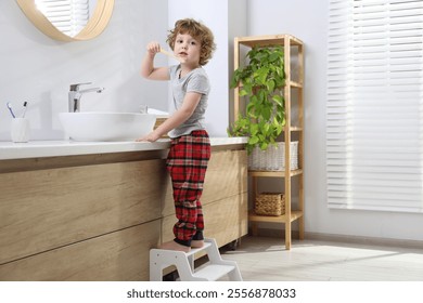 Little boy brushing teeth while standing on step stool near bathroom vanity indoors - Powered by Shutterstock