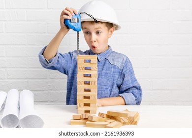 Little boy builder engineer in white construction helmet plays and builds high skyscraper from wooden blocks or cubes and plans. Emotional surprised child measures the tower with a measuring tape. - Shutterstock ID 2161936393