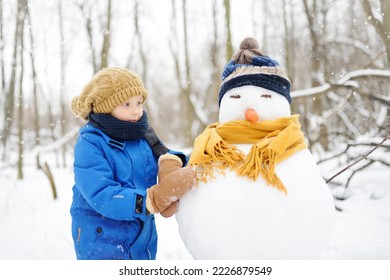 Little boy building snowman in snowy park. Child embracing snowman wearing hat and scarf. Active outdoors leisure with family with children in winter. Kid during stroll in a snowy winter park